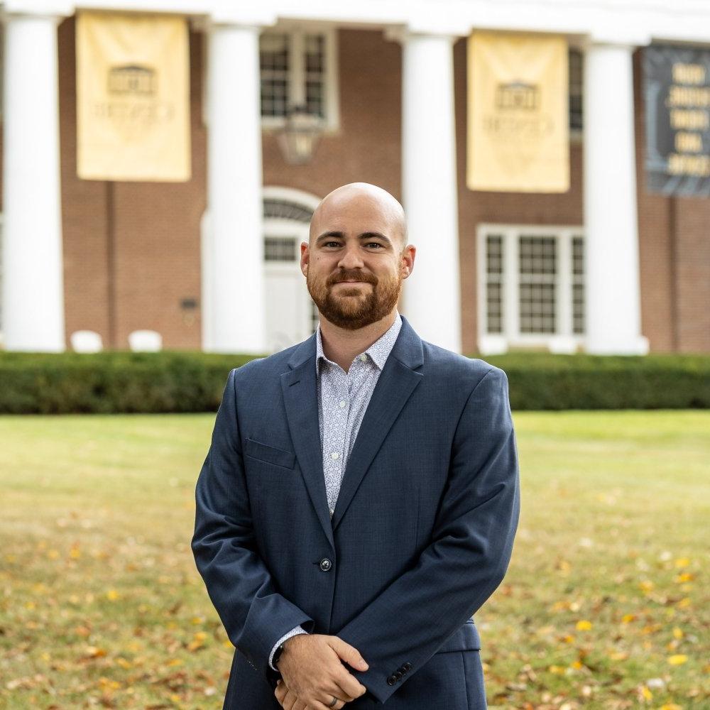 Man with facial hair wearing sports jacket with plaid dress shirt in front of Old Centre