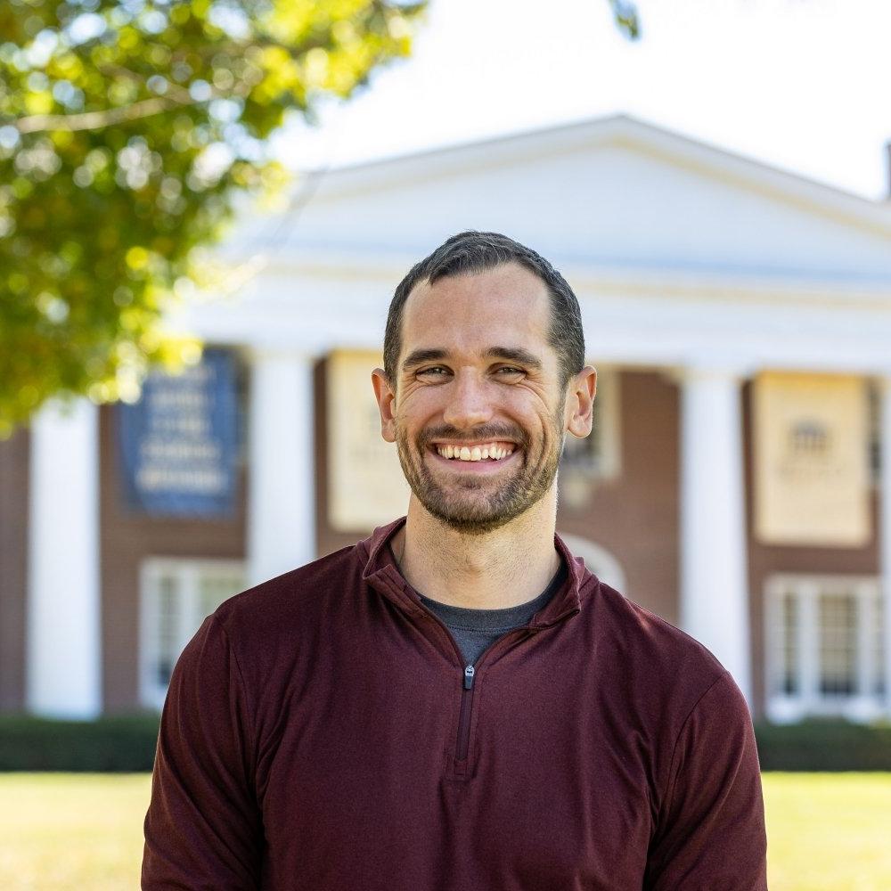 Man with dark hair and beard wearing maroon shirt in front of Old Centre